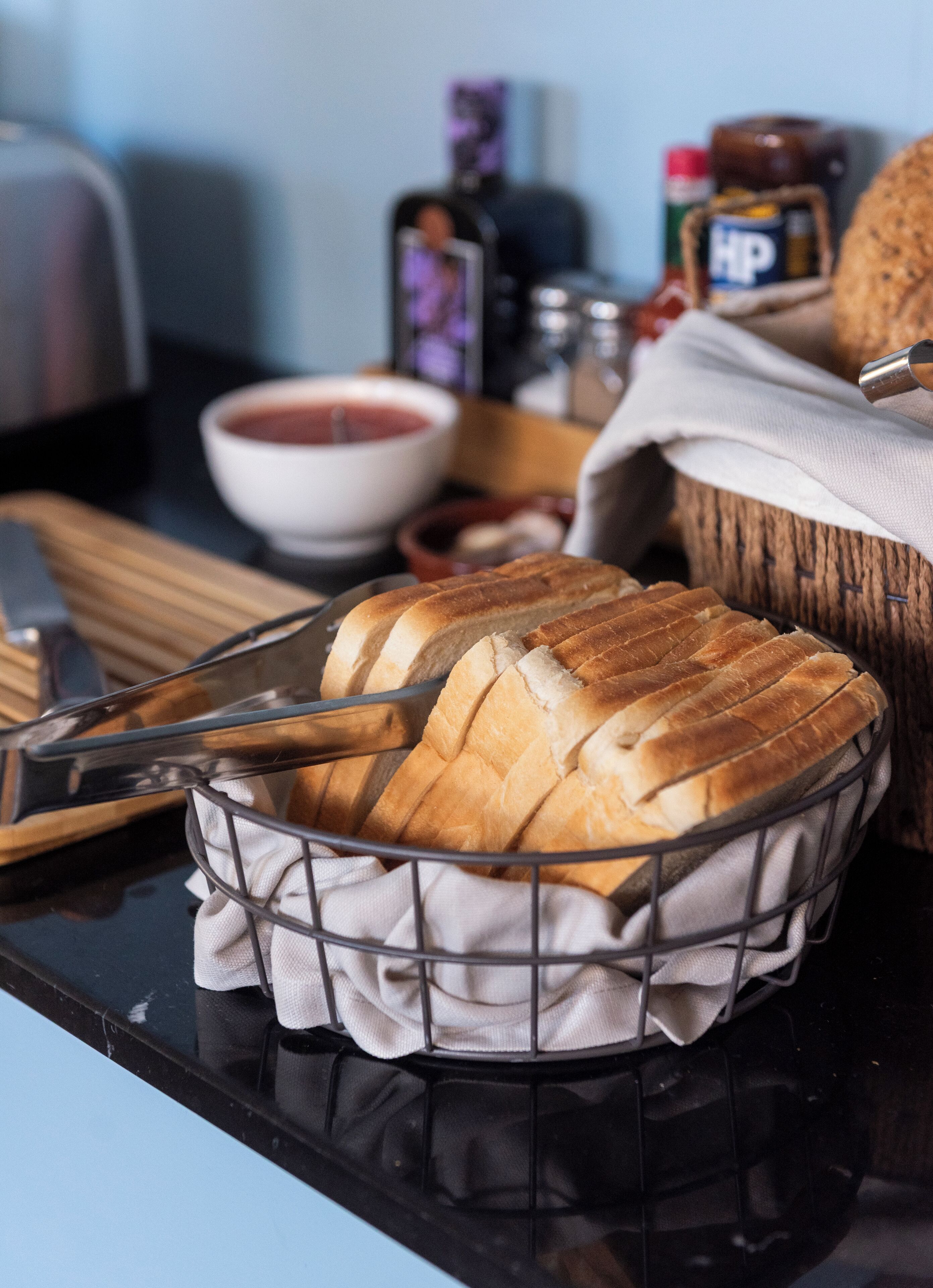 A basket of sliced bread on a kitchen counter with a knife, a bowl of soup, and condiments.