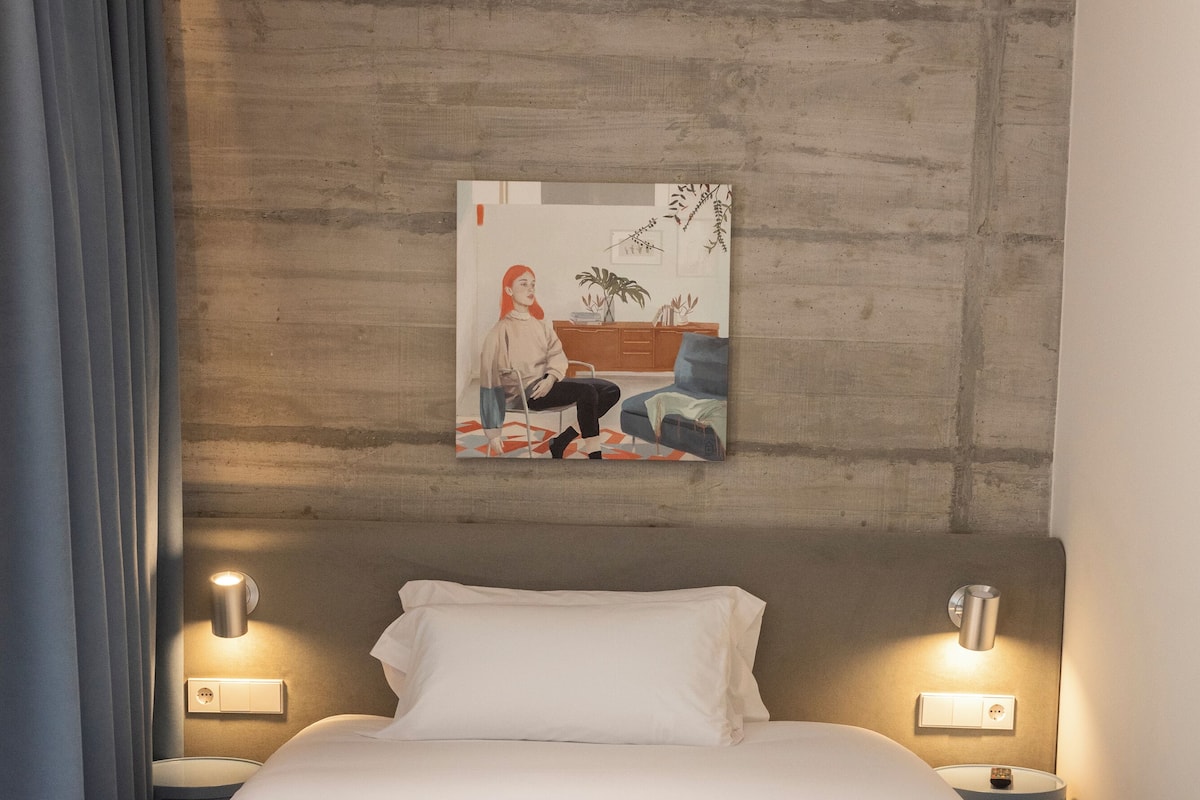Un lit bien fait, avec une serviette blanche roulée, deux tables de chevet avec des lampes, un tableau au mur et une tête de lit en bois.