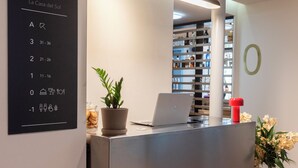 Un mostrador de recepción con una computadora portátil, una planta en maceta y un objeto rojo encima. En la pared, hay un cartel con números de habitación y símbolos.