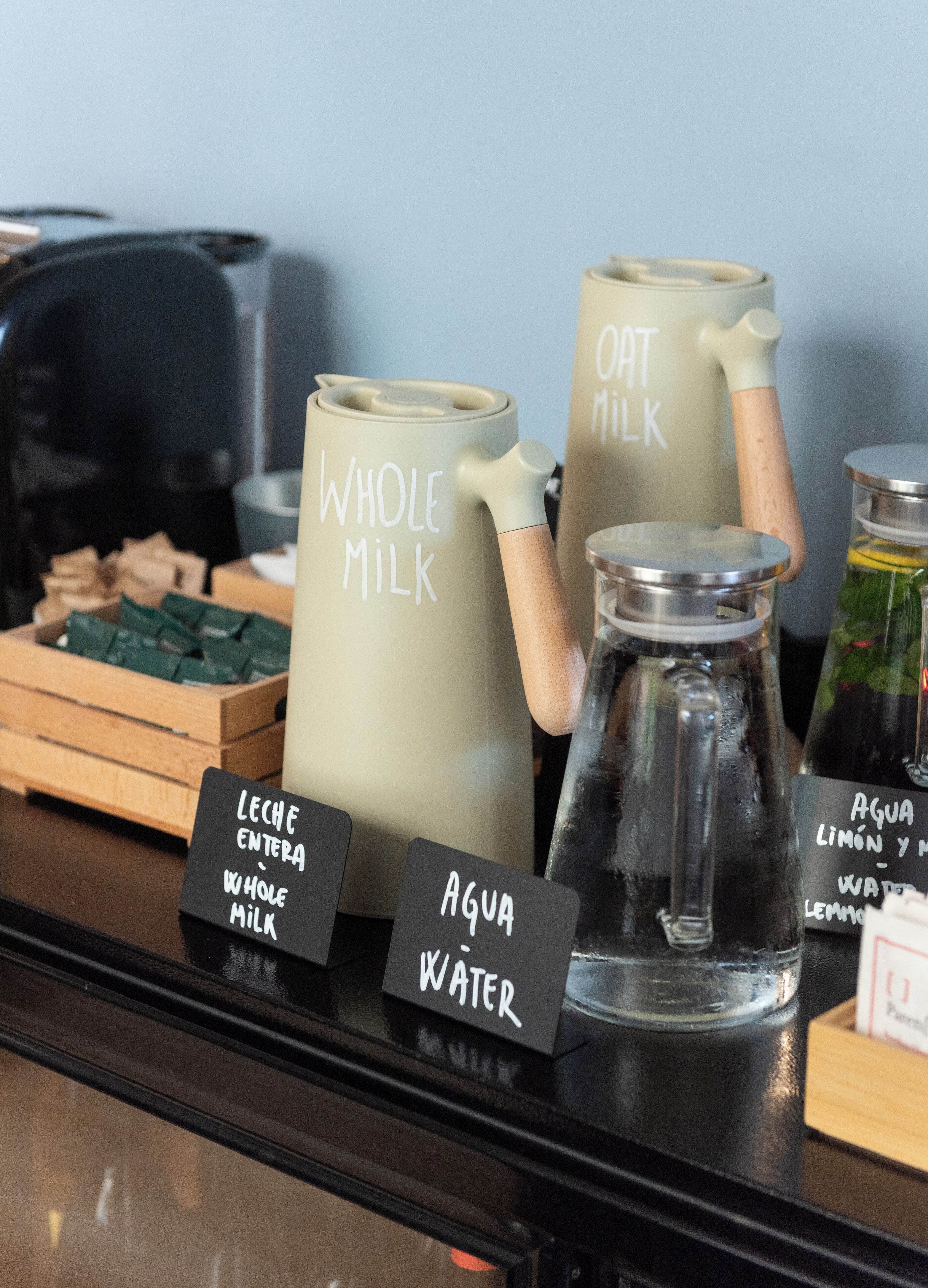Una estación de café con dispensadores etiquetados para leche entera, leche de avena y agua.
