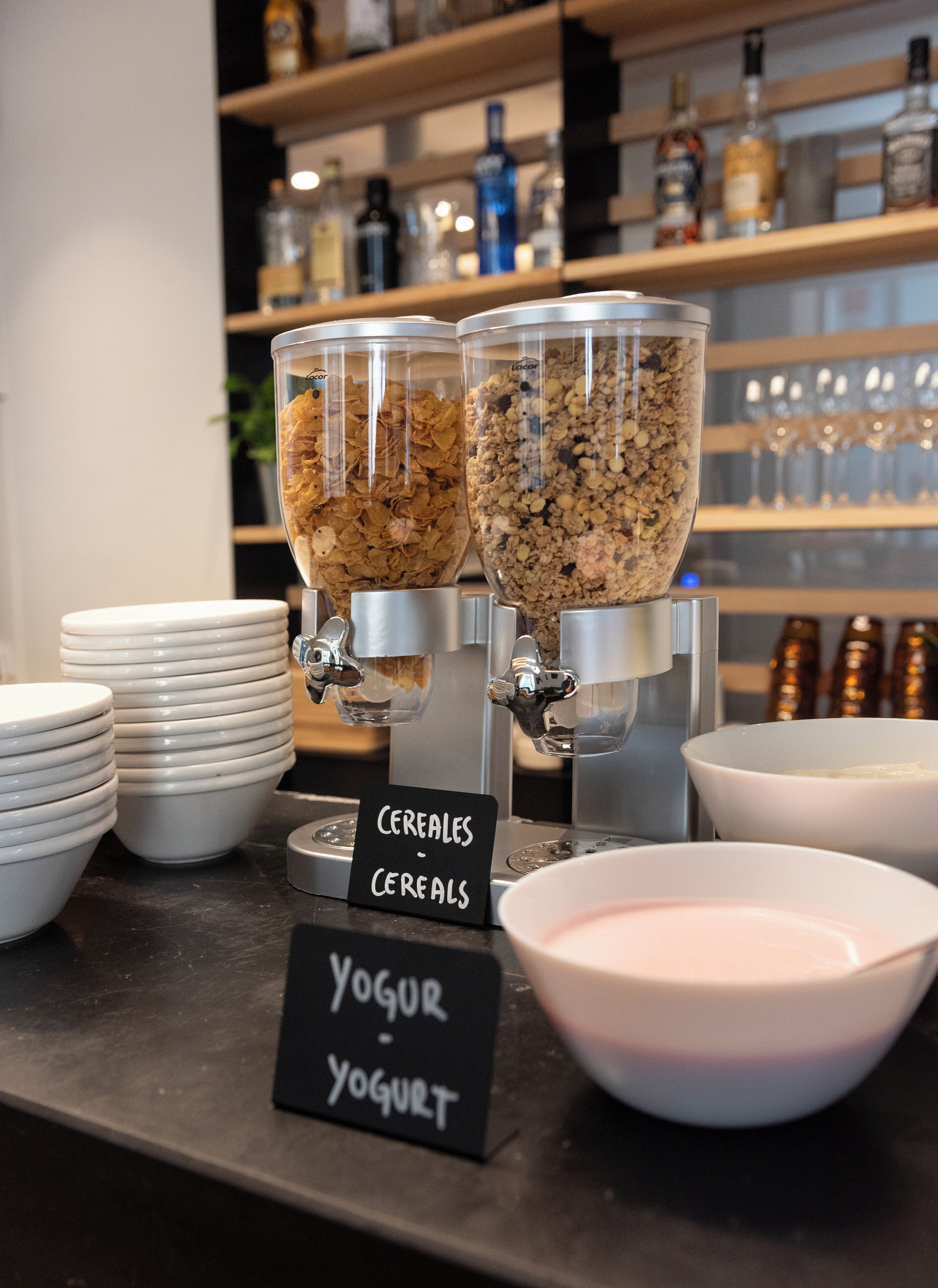 Una barra de desayuno con dispensadores de cereales y yogur, platos blancos y un cartel que indica las opciones de comida.