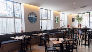 Un restaurante moderno con mesas de madera y sillas negras, ventanas amplias con cortinas y una zona de estar acogedora.