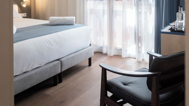Una habitación de hotel con una cama, una silla, una mesita de noche y un ventanal con cortinas.