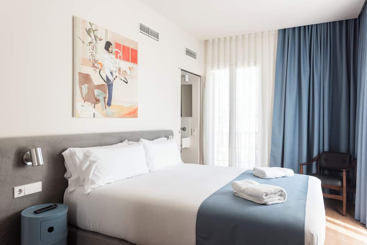 Une chambre d’hôtel avec un lit, une chaise, un tableau et une fenêtre avec des rideaux.