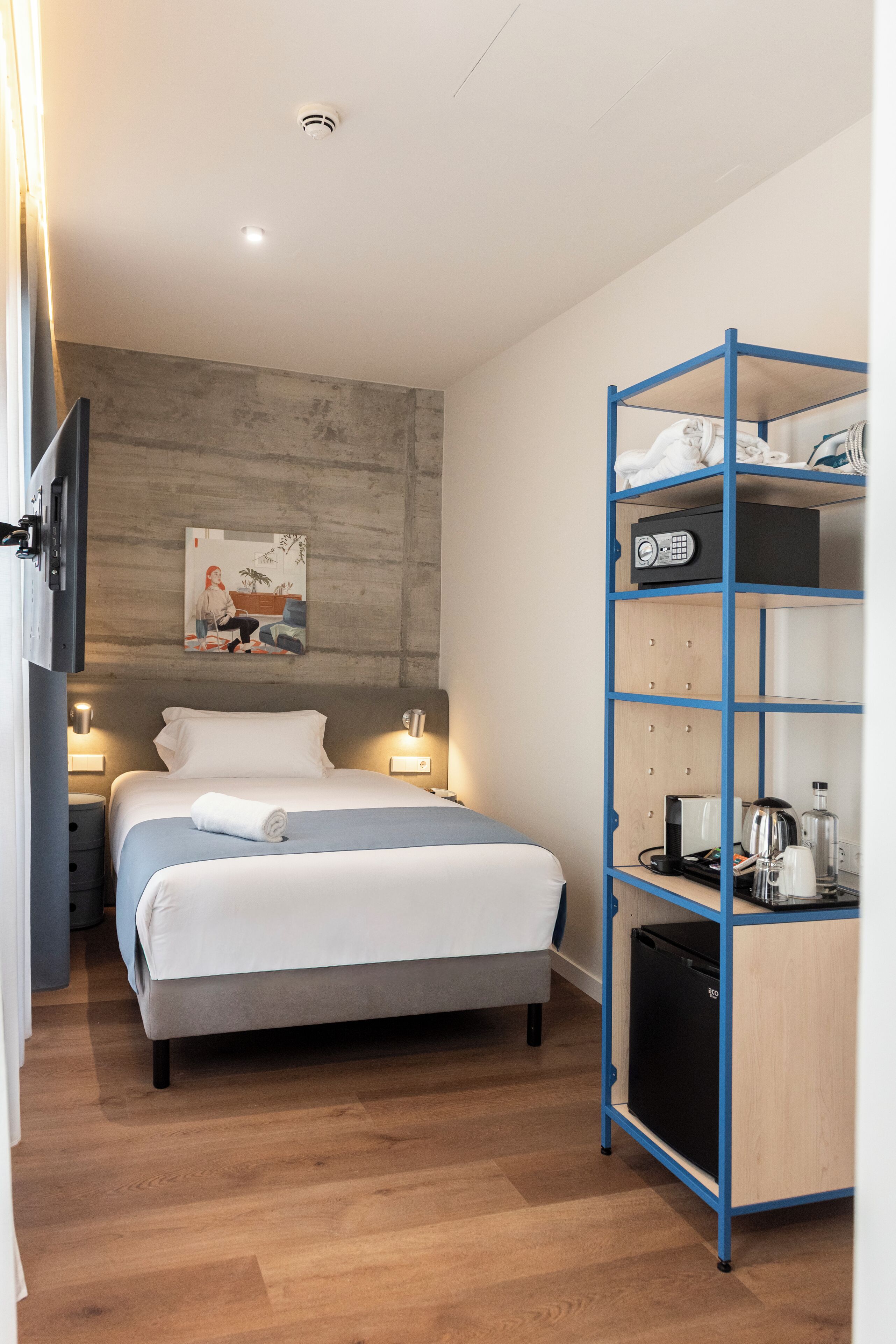 A hotel room with a bed, bedside lamps, a wall-mounted artwork, a blue shelving unit with a kettle and other items, and a small black appliance.