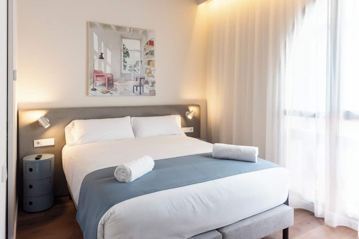 Un lit bien fait, avec du linge de lit blanc et un élément décoratif bleu, une lampe de chevet et une petite table de chevet avec une télécommande.
