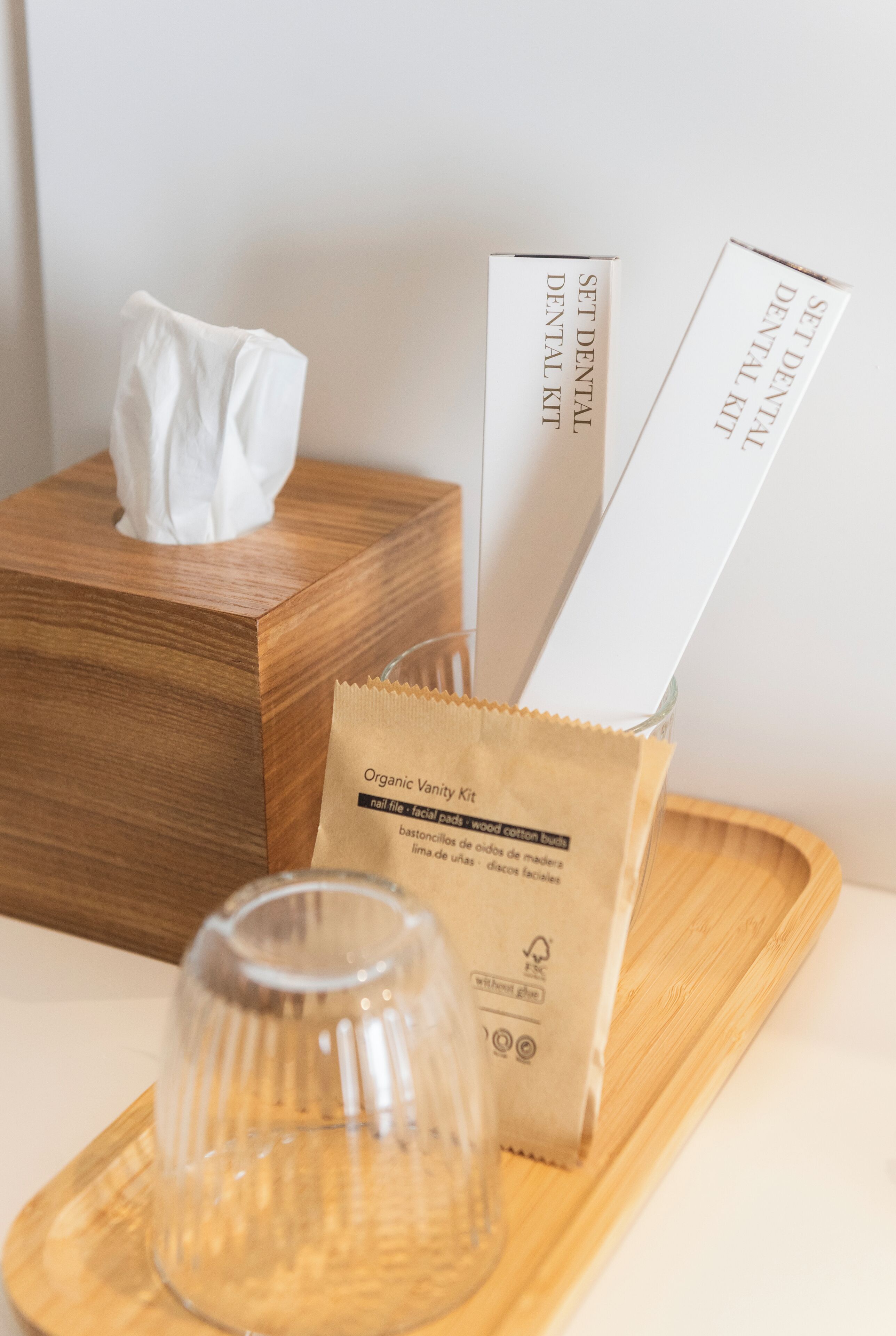 Una bandeja de madera con una caja de pañuelos, un set de productos para la higiene bucal y un recipiente de vidrio.