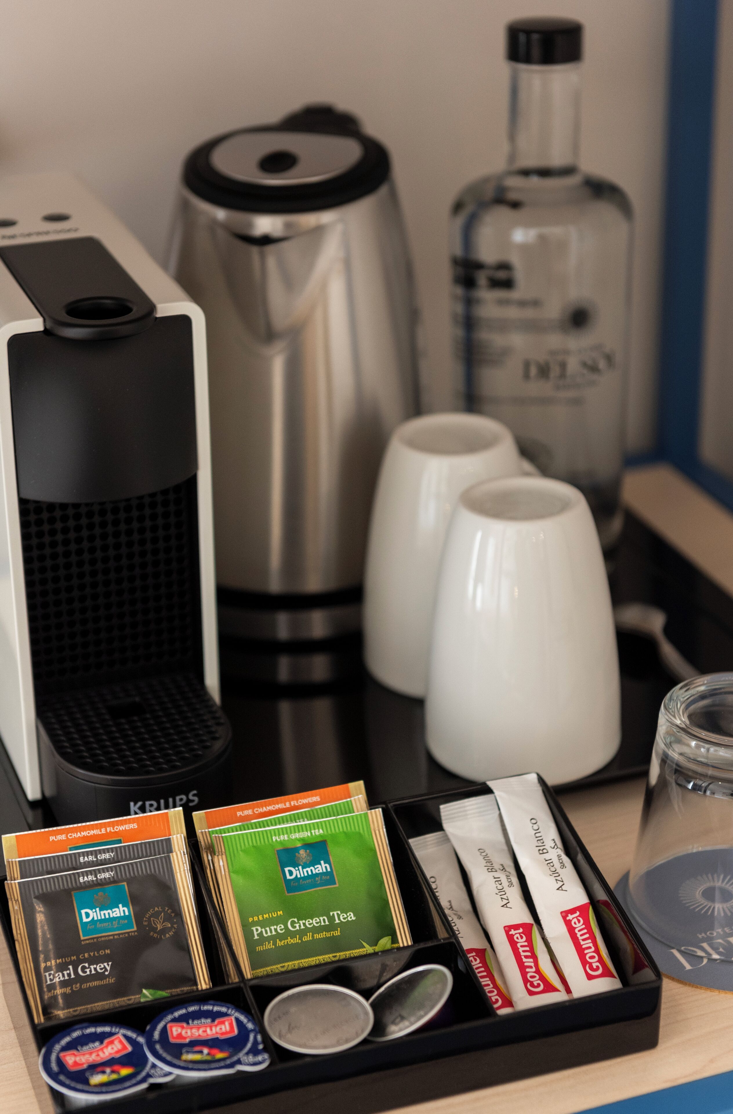 Una cafetera, un hervidor, dos tazas blancas, una botella de licor y cajas de té sobre una mesa.