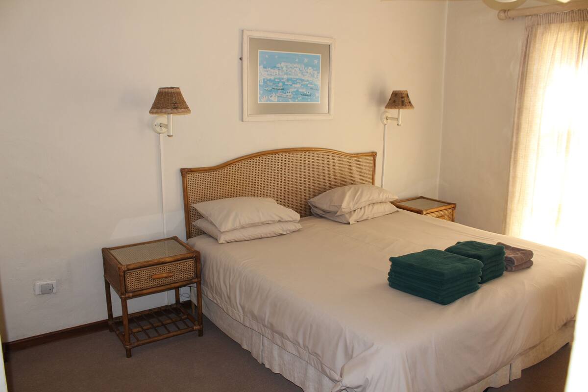 Una cama bien tendida con ropa de cama blanca y dos almohadas, flanqueada por dos mesitas de noche de mimbre con lámparas, en una habitación con pared blanca lisa y un cuadro enmarcado sobre la cama.