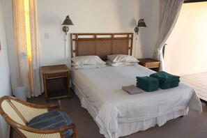 A neatly made bed with white linens, two bedside tables with lamps, a wicker chair, and a view of the outdoors through a window with curtains.