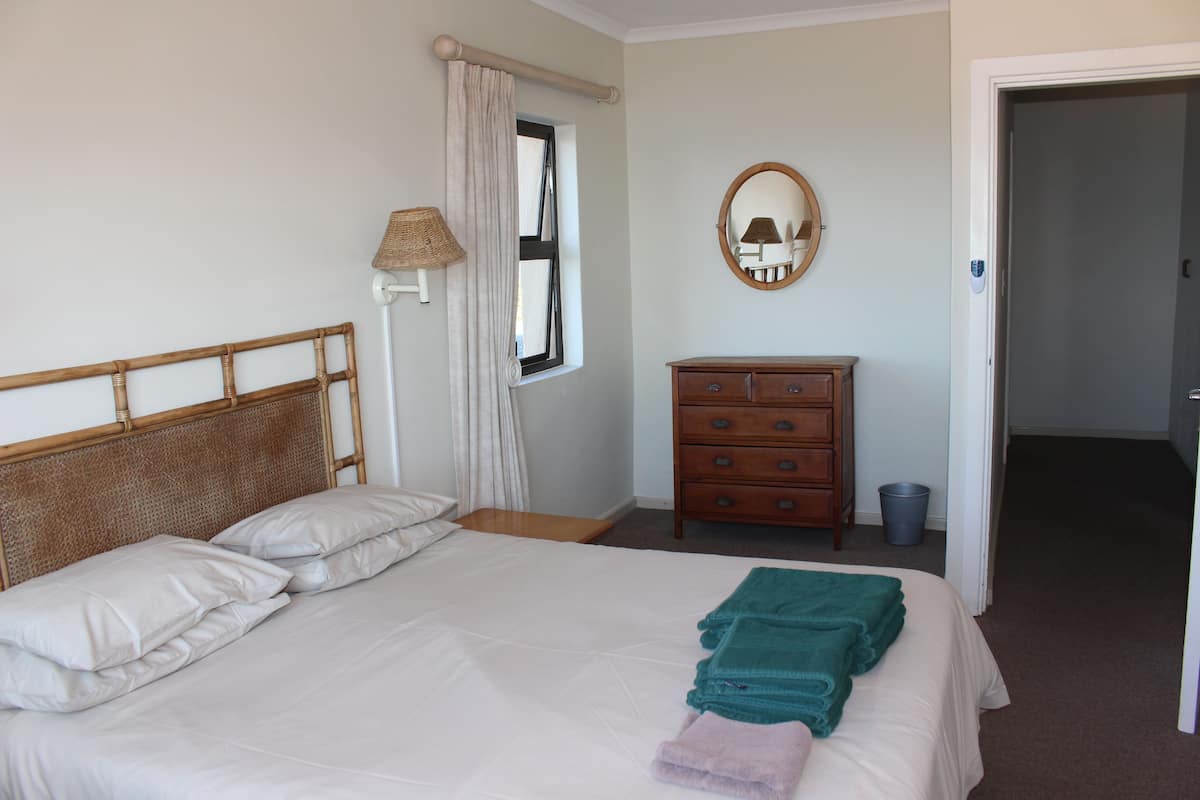 Una cama bien hecha con almohadas y toallas dobladas, un aparador de madera, un espejo y una ventana con cortinas.