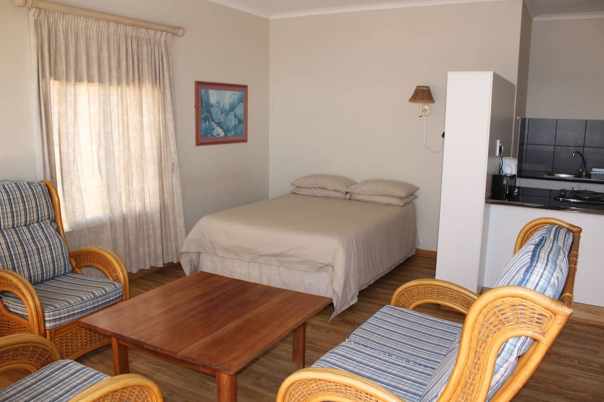 Habitación pequeña con cama, una mesa de madera, sillas de mimbre, una kitchenette y un cuadro en la pared.