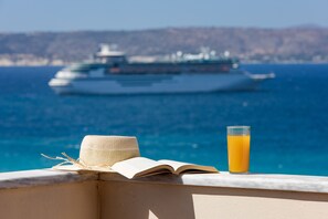 Un balcón con un sombrero de paja, un libro abierto y un vaso de jugo de naranja. Al fondo, se divisa un crucero en el agua.