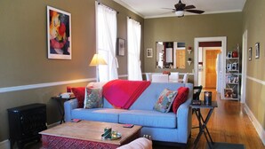 Un salon avec un canapé bleu, une table basse en bois et un plaid rouge. On y trouve une lampe sur une table d’appoint, une bibliothèque et une porte qui donne sur la cuisine.