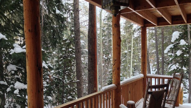 A wooden deck with a railing, a rocking chair, and a bench covered in snow, surrounded by snow-covered trees. - Forest Creek Cabin 3 Bedroom Holiday Home By Pinon Vacation Rentals (Monarch)