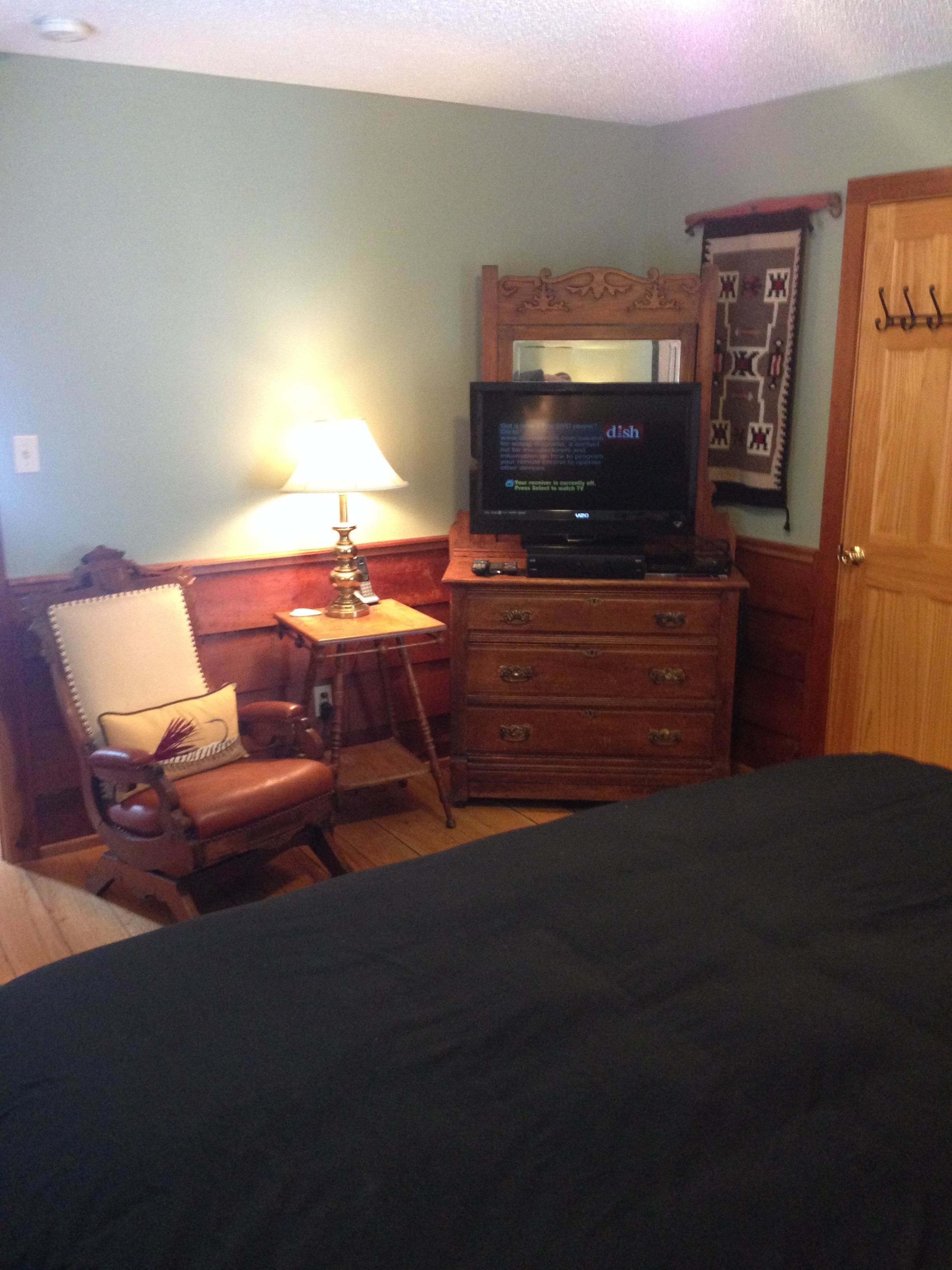 A hotel room with a bed, a television on a wooden dresser, a chair with a cushion, a lamp, and a wooden door.