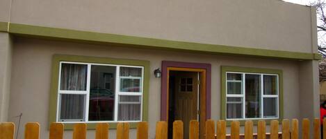 A two-story building with a beige exterior and green trim. There are two windows on the ground floor, one with a purple door, and a wooden fence in the foreground.