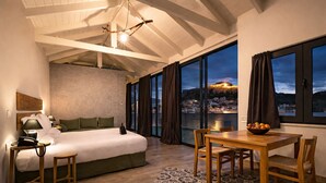 A bedroom with a bed, bedside table, lamp, and a view of a cityscape at dusk.