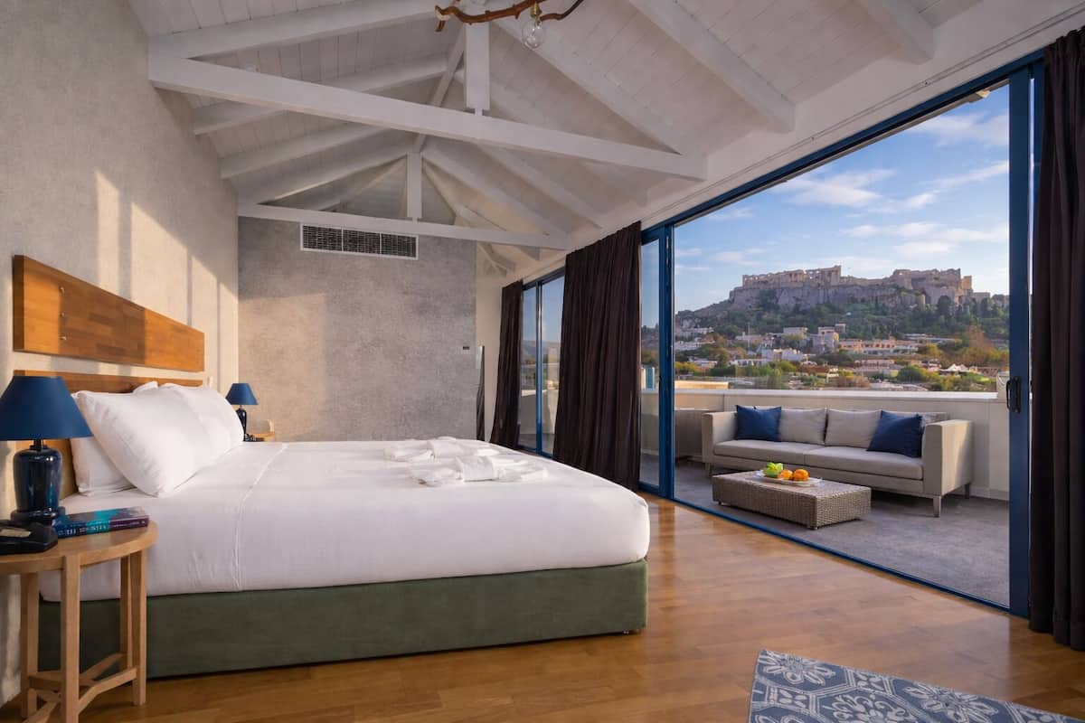 A bedroom with a large bed, a nightstand, a lamp, a view of a cityscape through a large window, and a balcony with a sofa and table.