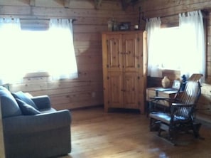 Ein gemütliches Blockhaus-Interieur mit Holzboden, einer Couch, einem Esstisch mit Stühlen und einem Schrank.
