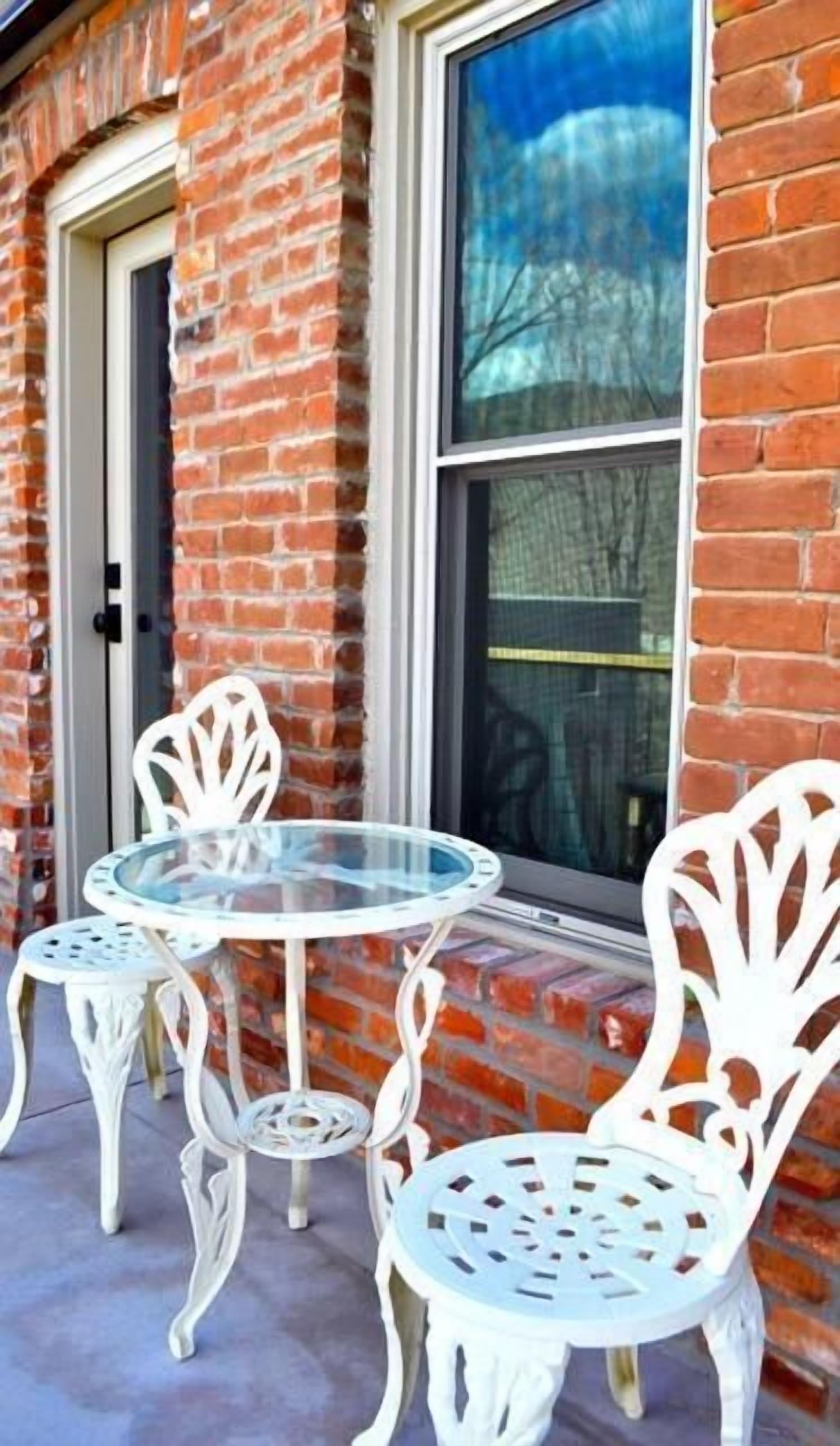 A round glass table with four white metal chairs, positioned outside a brick building with a window and a door.