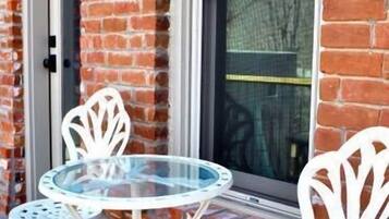 A round glass table with four white metal chairs, positioned outside a brick building with a window and a door.