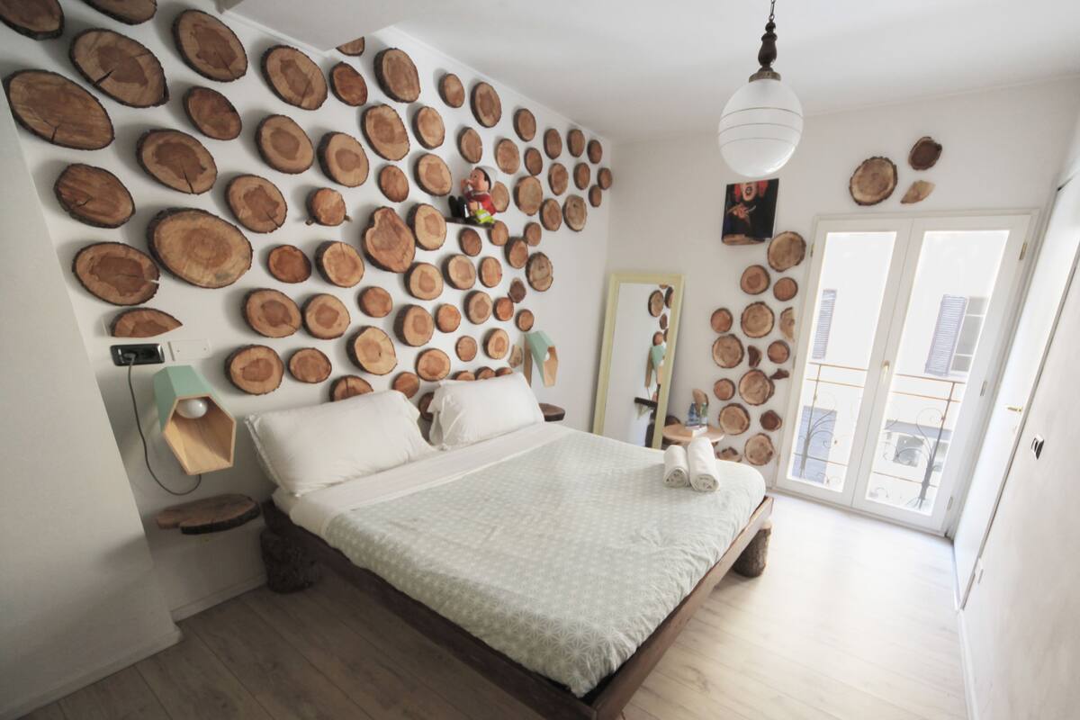 A bedroom with a bed centered on a wooden floor, surrounded by a wall of circular wooden slices. A hanging light fixture and a mirror are visible.
