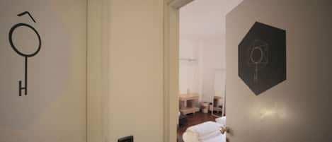 Habitación de hotel con cama, un pequeño escritorio y una silla. Se observa una puerta con el símbolo de un ojo de cerradura.