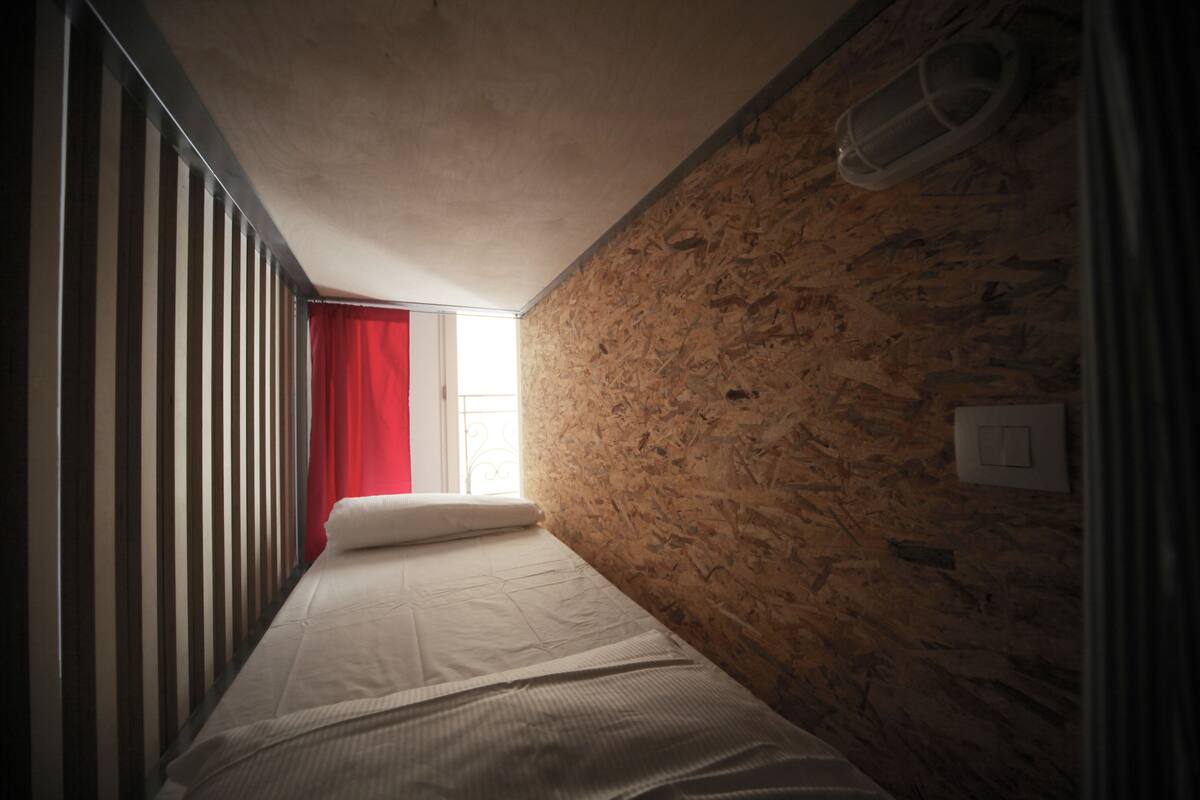 A narrow room with a sloped ceiling, a bed with white bedding, a red curtain, and a window with a view of the outside.