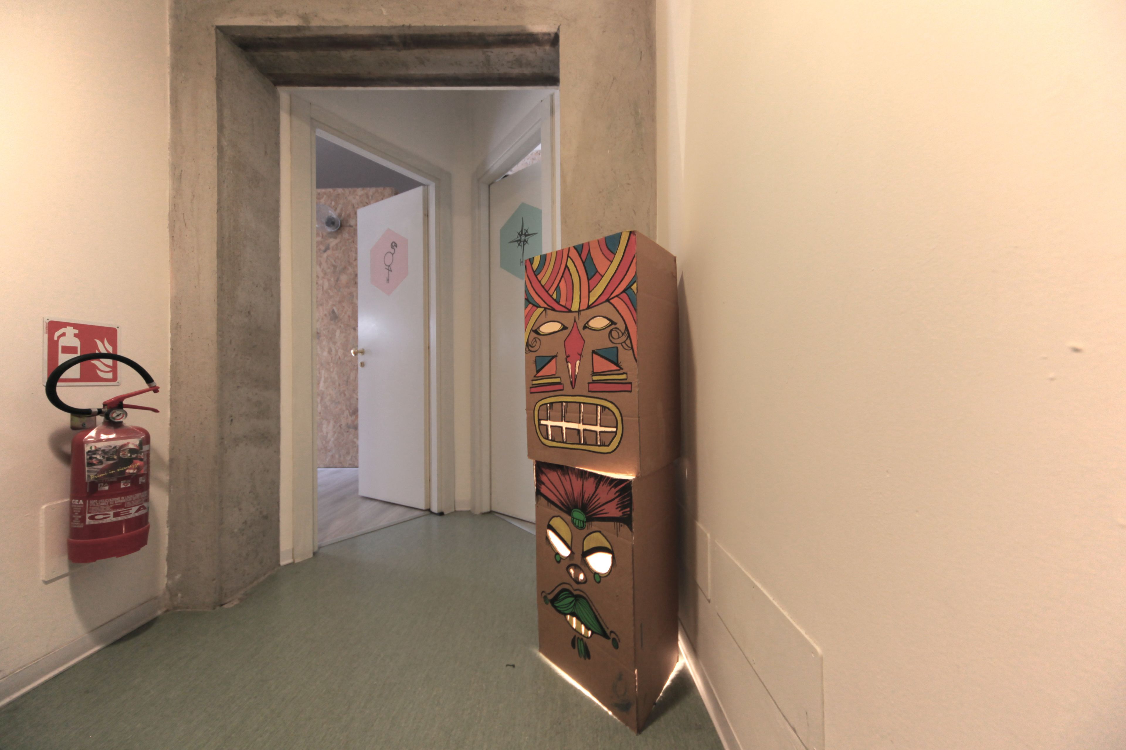 Un pasillo con un extintor a la izquierda, una puerta al final y un objeto decorativo con rostro a la derecha.