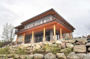 A modern two-story house with a stone foundation, large windows, and a covered porch supported by wooden pillars.