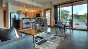 A spacious living area with a sofa, coffee table, and dining set. A kitchen with wooden cabinets and a refrigerator is visible. Large windows offer a view of the outdoors. - River Time 1 Bedroom Holiday Home By Pinon Vacation Rentals (Salida)
