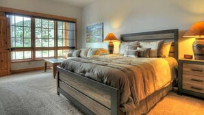 A bedroom with a large bed, two bedside tables, a nightstand, and a window with a view of greenery. - River Time 1 Bedroom Holiday Home By Pinon Vacation Rentals (Salida)