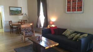 A living room with a dark blue sofa, a wooden coffee table, a floral armchair, a dining table with chairs, a painting, and a bird-themed artwork.