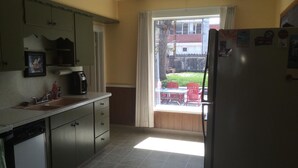 A kitchen with a sink, cabinets, and a refrigerator. There is a window with curtains and a view of a backyard with a table and chairs.