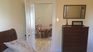 A bedroom with a bed, a dresser, a mirror, and a dining area visible through an open door.