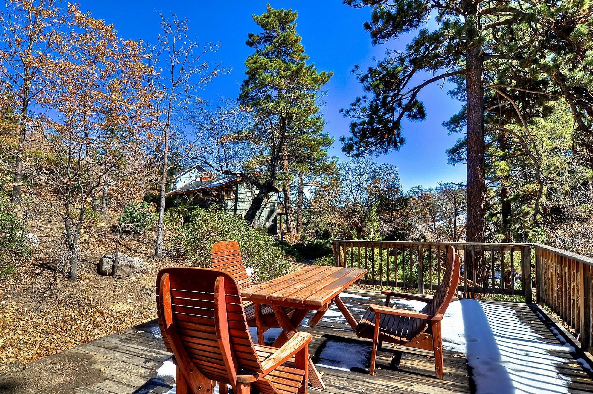 A wooden deck with a picnic table and chairs, surrounded by trees and a house in the background.