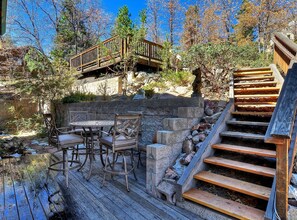 Una terraza de madera con mobiliario exterior, un muro de piedra y una escalera de madera que lleva a otra terraza.
