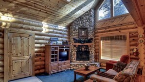 A log cabin interior with a stone fireplace, wooden furniture, and a large window.