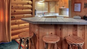 A log cabin interior with a kitchen area featuring a bar and stools, wooden walls, and a window with blinds.