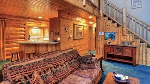 A log cabin interior with a wooden staircase, a sofa with patterned cushions, a bar area, and framed pictures on the walls.