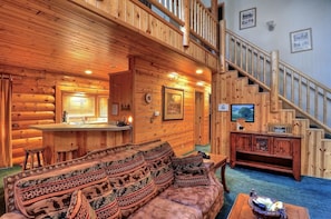 Interior de cabana de madeira com escada, sofá com almofadas estampadas, área de bar e quadros nas paredes.