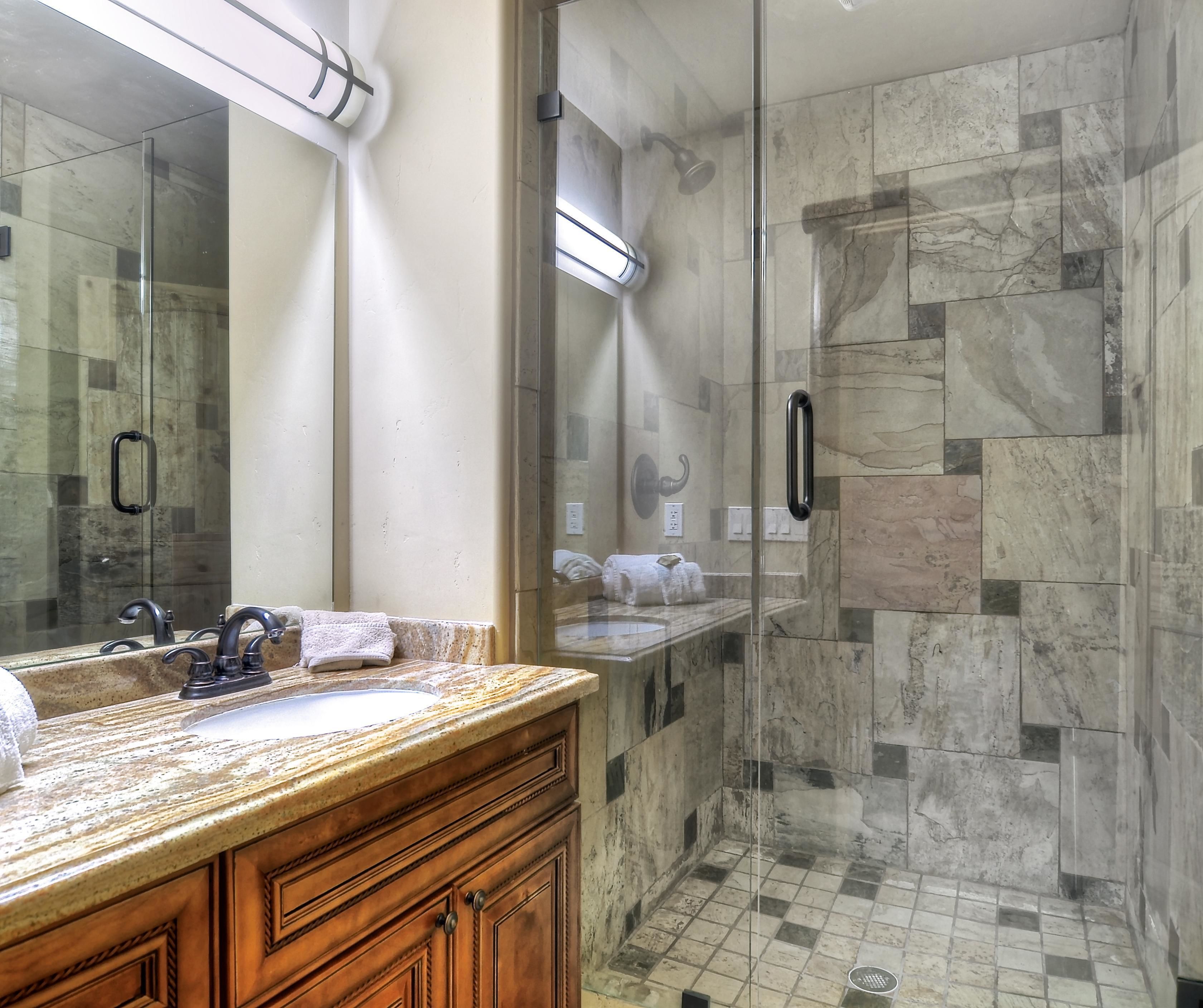 A modern bathroom with a granite countertop, a large mirror, a glass-enclosed shower, and marble tiles.