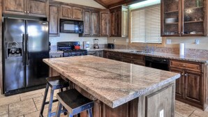 A kitchen with wooden cabinets, a stainless steel refrigerator, a granite countertop, and a window with blinds.