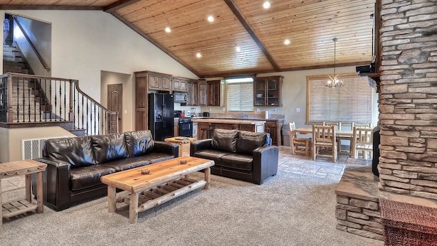 A spacious living area with a high ceiling, wooden beams, a stone fireplace, and a kitchen in the background.