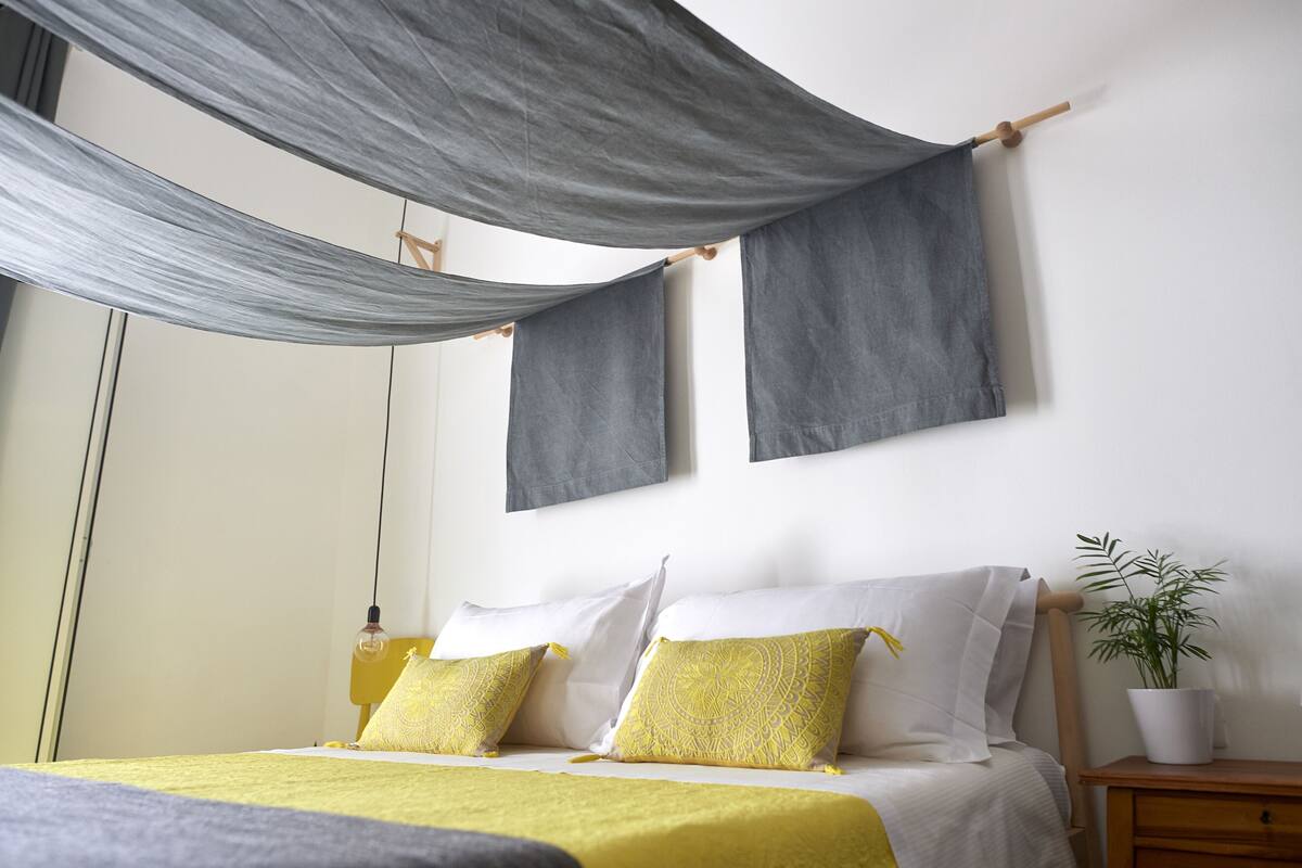 Ein Bett mit einer gelb-weißen Tagesdecke, zwei Kissen und einem grauen Stoffvorhang darüber.