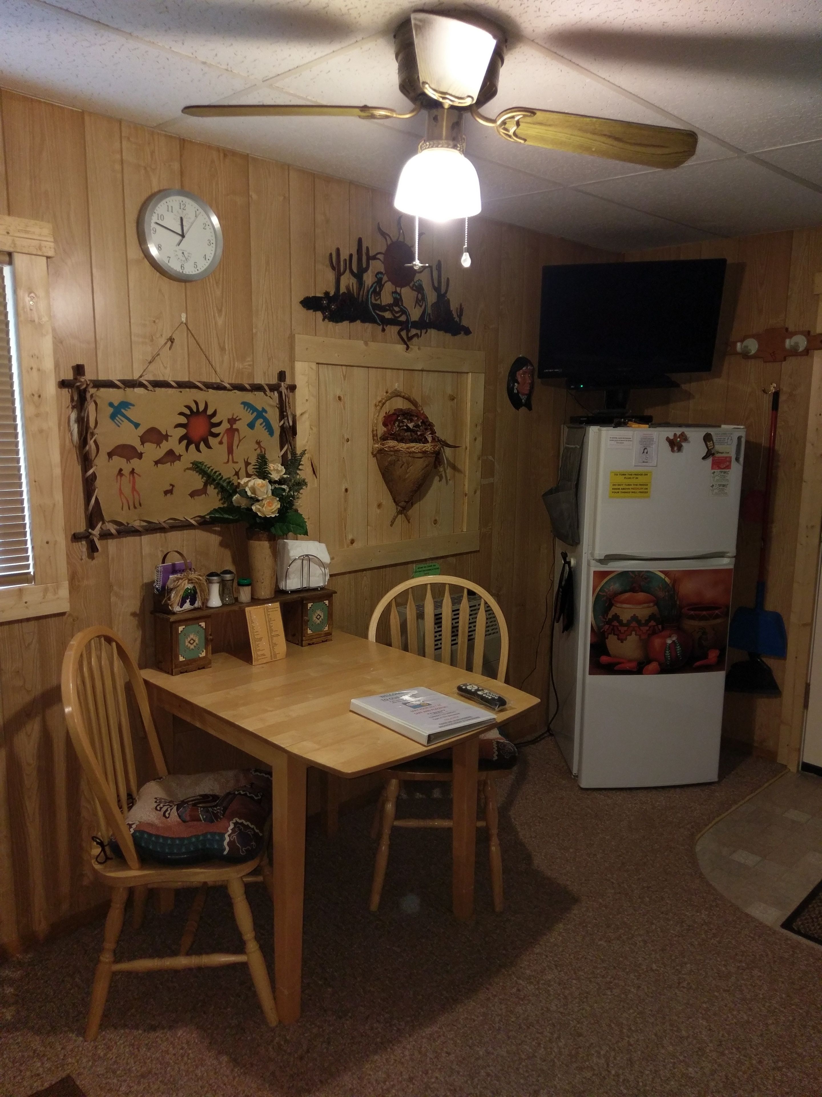 A wooden-paneled room with a dining table, chairs, a clock, a ceiling fan, a refrigerator, and a television.