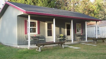 A small, single-story building with a porch, two windows, and a bench.