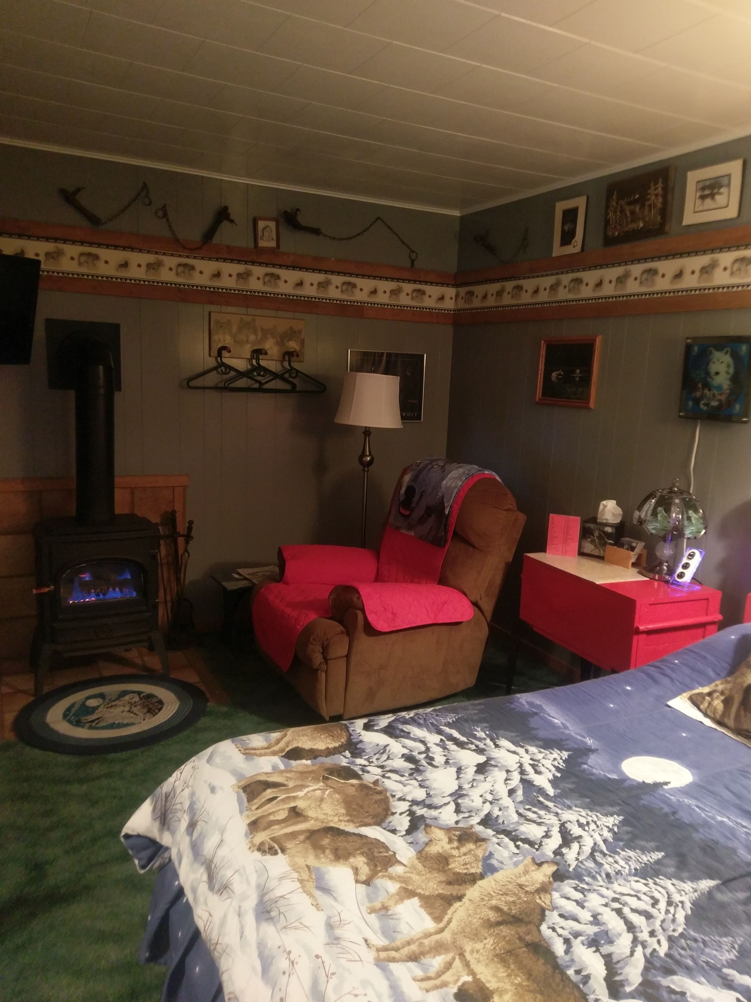 A cozy room with a fireplace, a red armchair, a desk with a lamp, and a bed with a wolf-themed blanket.