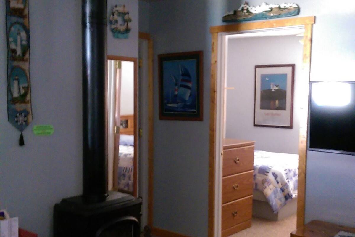 Un salon chaleureux avec un poêle à bois, un lit recouvert d’une couverture à rayures bleues et blanches, et une commode.